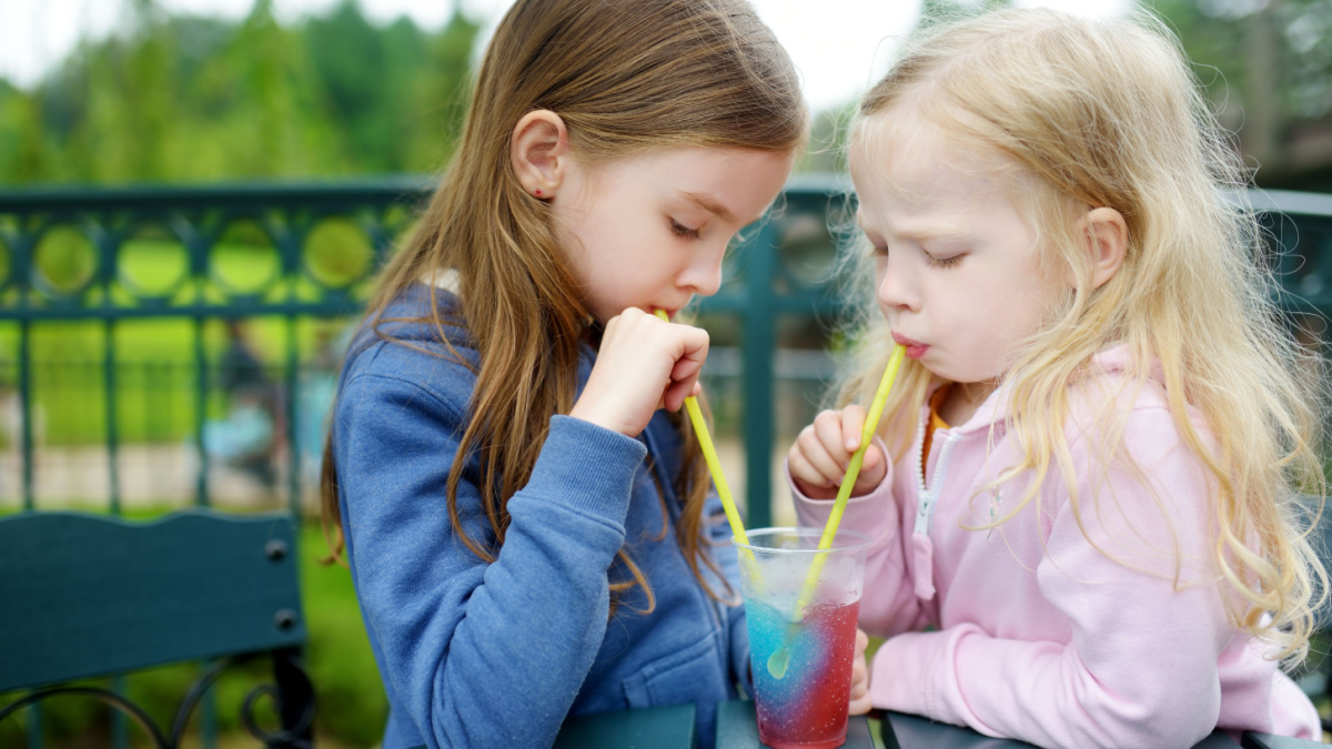 Two cute little sisters drinking colorful frozen slushie drink on hot summer day