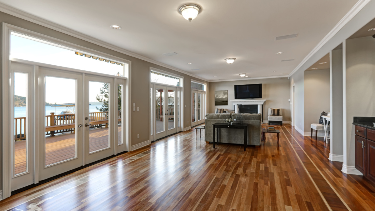Luxury spacious family room interior with wall of glass doors leading out to spacious deck and facing the lake, polished hardwood floor and cozy sitting area with fireplace .