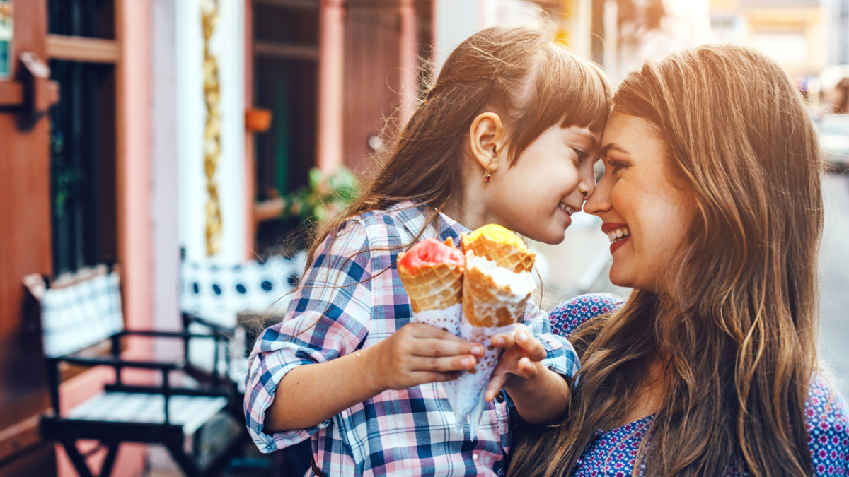 Mom with her 6 years old daughter walking along city street and eating ice cream in front of the outdoor cafe. Good relations of parent and child. Happy moments together.