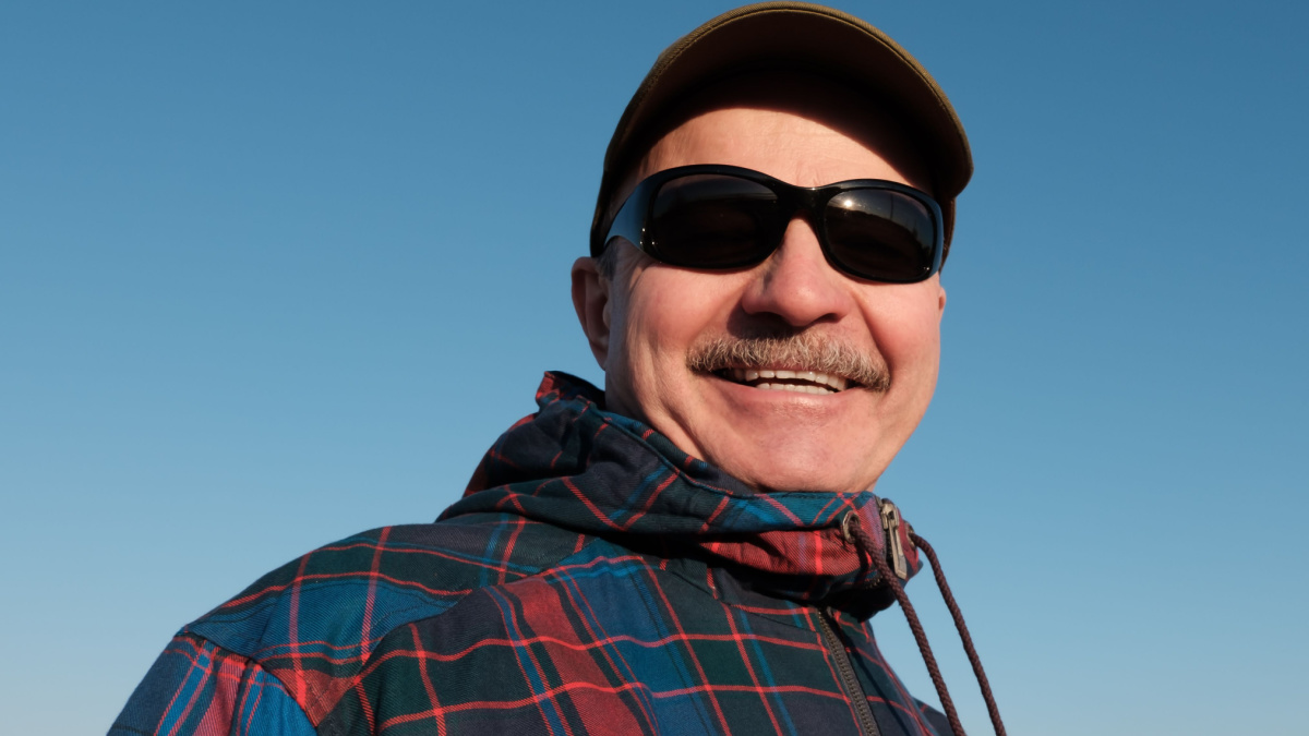 An elderly man in sunglasses and a baseball cap looks down and smiles.