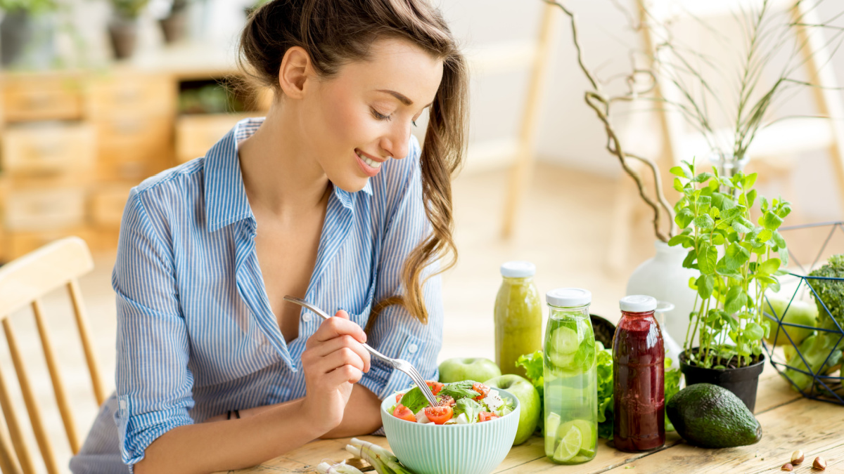 Young and happy woman eating healthy salad sitting on the table with green fresh ingredients indoors