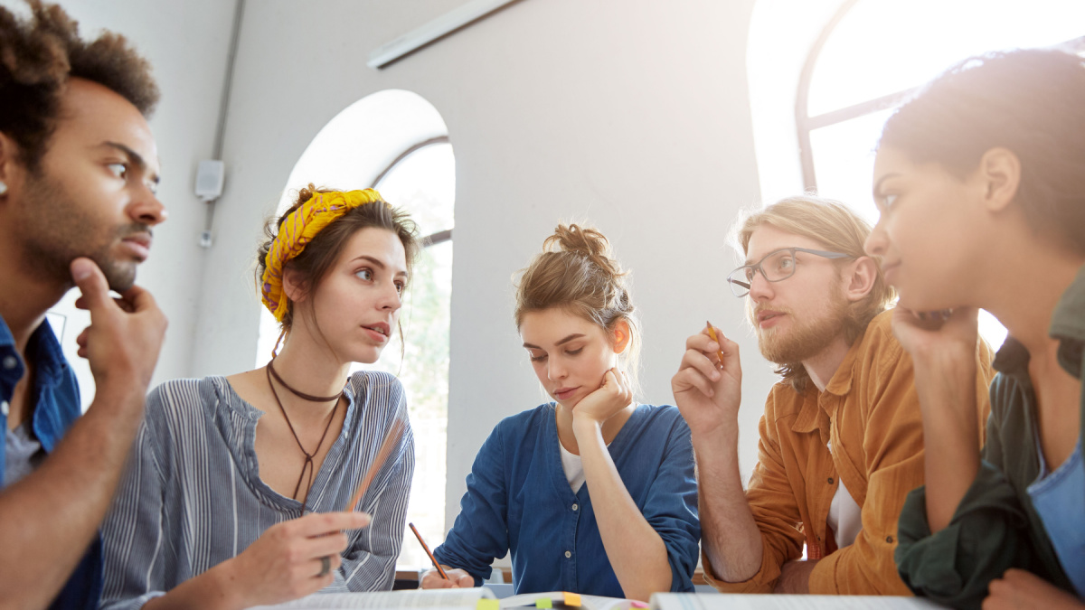 Collaboration and brainstroming concept. Portrait of interracial friends gathering together sitting at table in classroom surrounded with books having debates expressing their opinions and views.