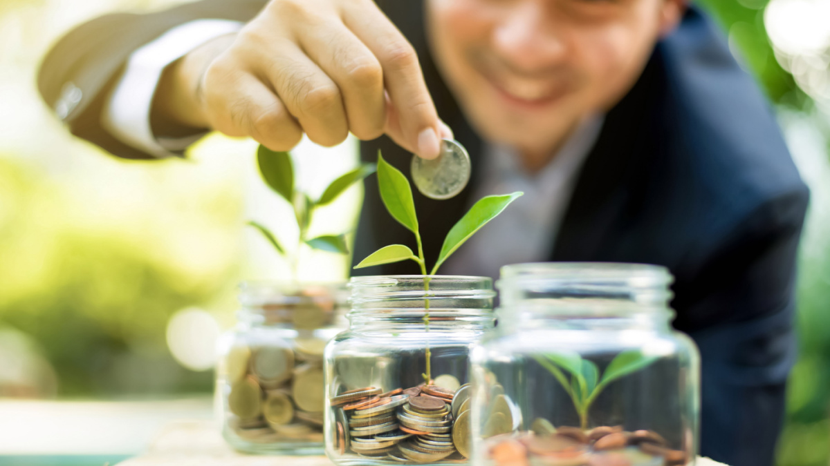 Businessman putting coin into the glass jar with young plant, demonstrating financial growth through saving plans and investment schemes.