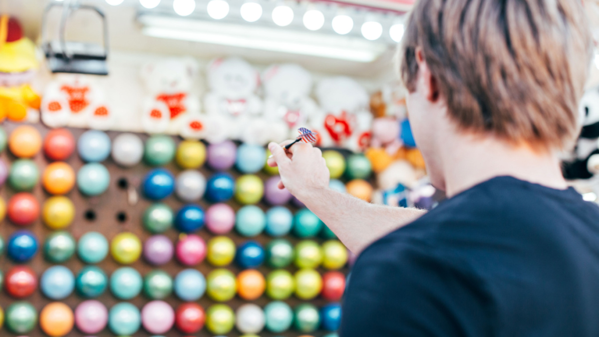 Young man aims at wall of colourful air balloons in order to win teddy bear or plush toy prize for his girlfriend, during romantic date at amusement park or carnival