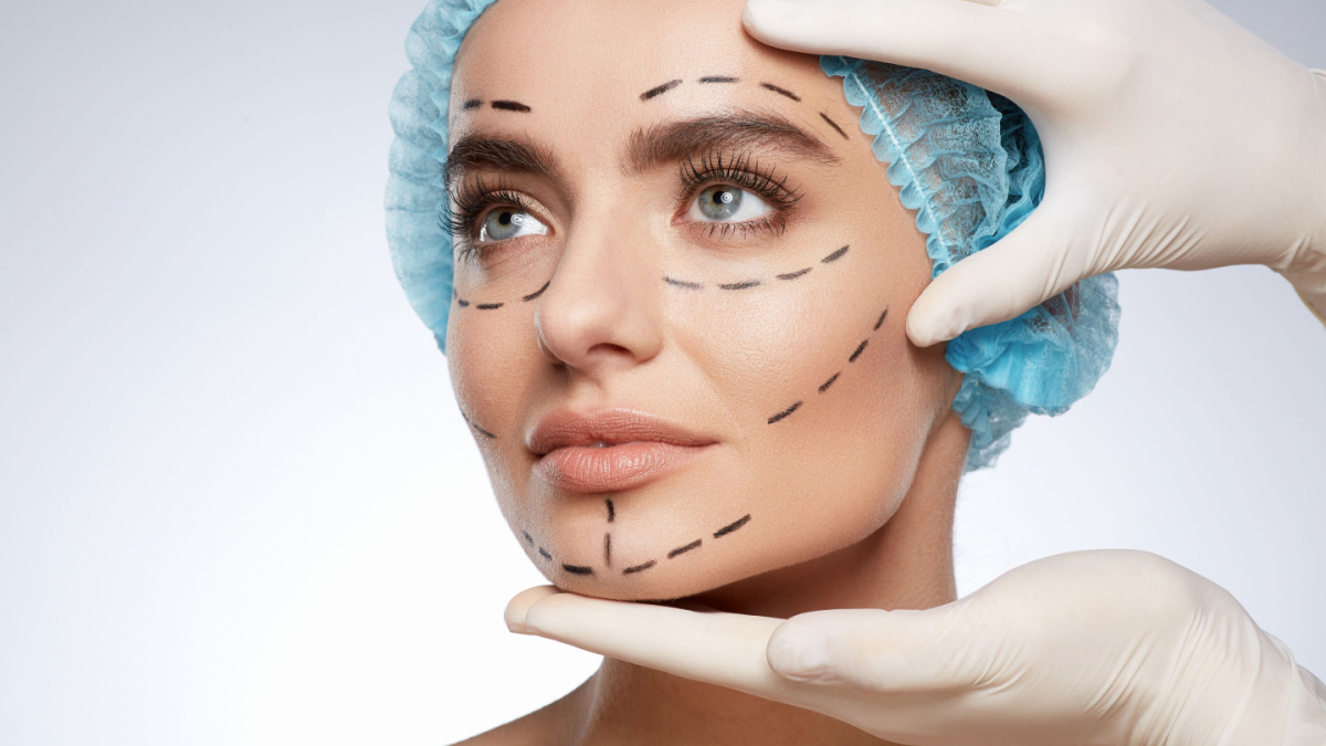 Beauty portrait of woman in blue cap looking aside, plastic surgery concept, studio. Hands in white gloves holding face of model with puncture lines, indoors