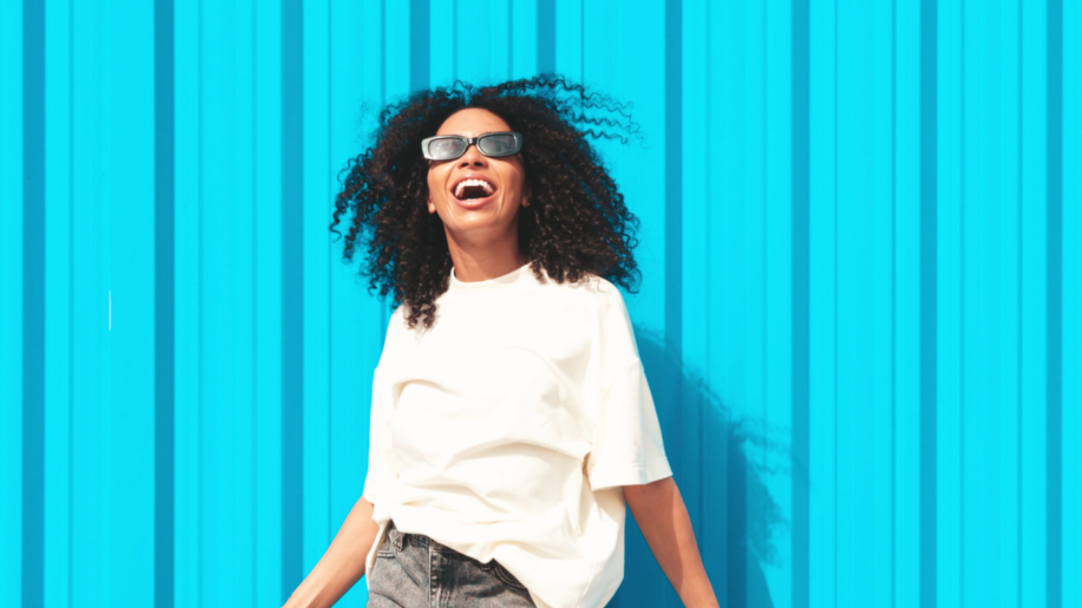 Beautiful black woman with afro curls hairstyle.Smiling hipster model in white t-shirt. Sexy carefree female posing in the street near blue wall in sunglasses.