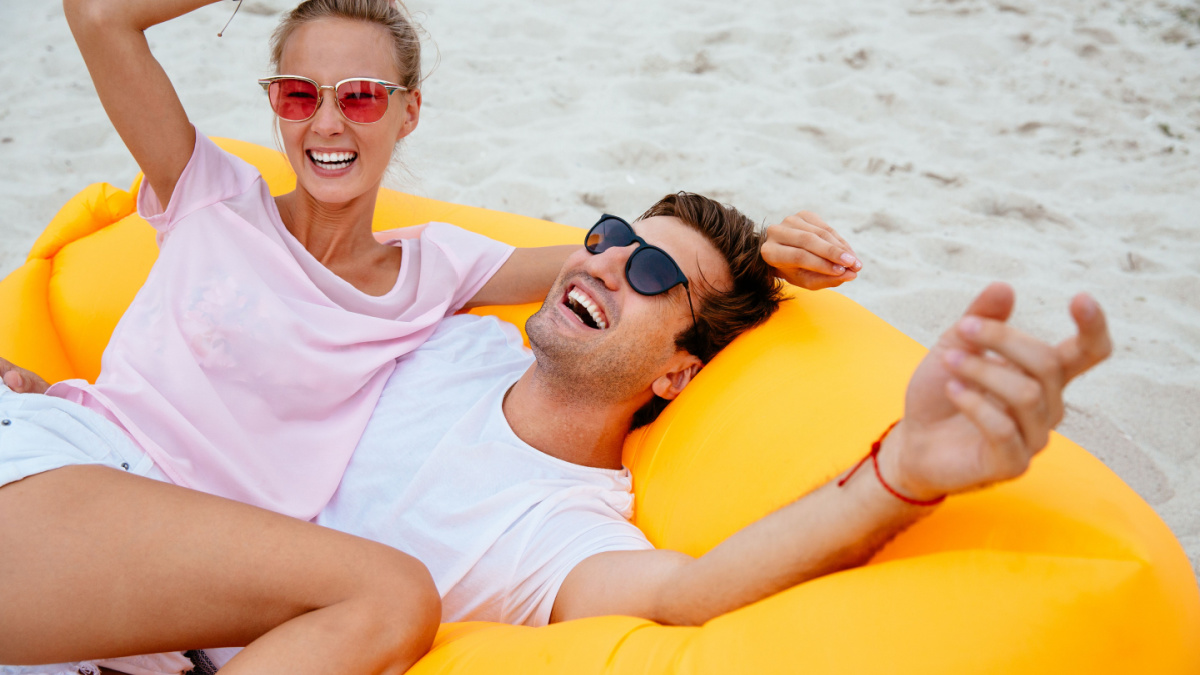 Attractive smiling guy and girl having fun together, laughing while laying on air sofa Lamzac. On the beach.
