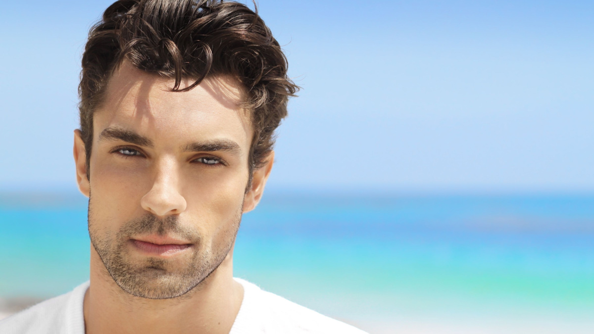 Handsome young man in casual white top against bright beach background.