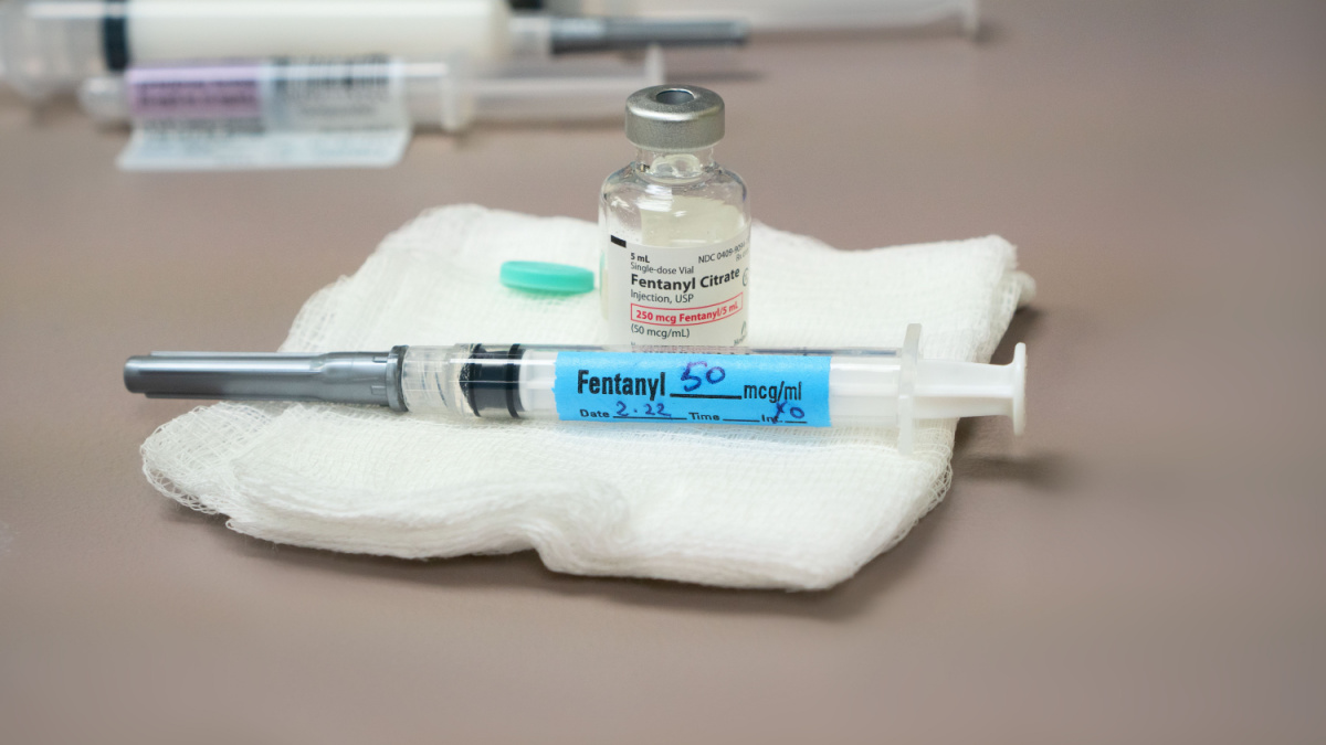 A 3 ml syringe labeled with a fentanyl sticker and an uncapped bottle of fentanyl, a potent intravenous opioid, on a top of an anesthesia cart.