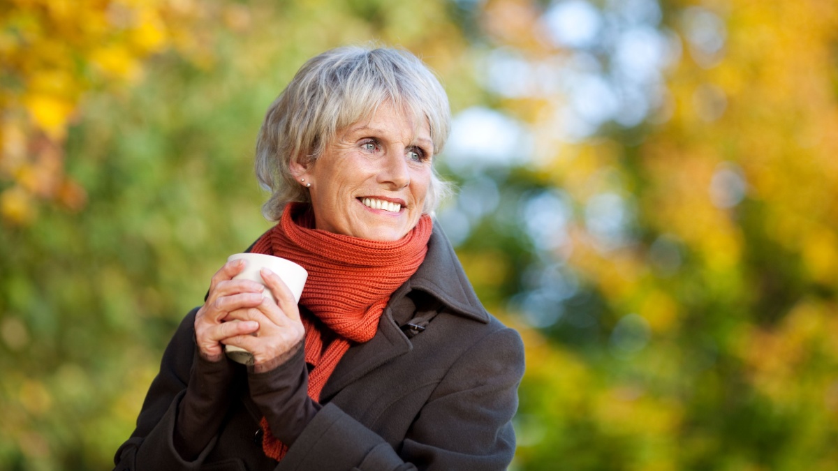 Smiling senior woman in jacket enjoying coffee in park.