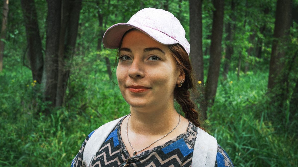 Young pretty woman traveller in baseball cap looks and smiles at camera at green nature forest background, portrait close up.