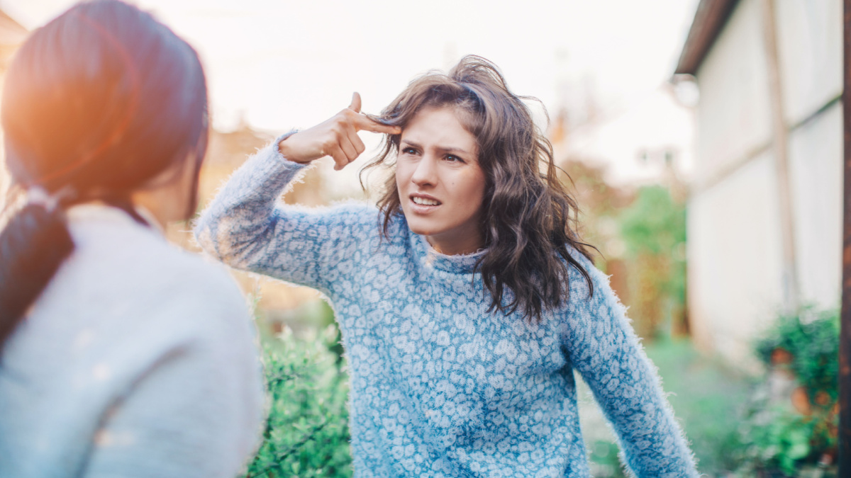 Two young women are cursing. Quarrel, aggression, stress. Two brunette girlfriends swear. Scream. Point fingers to the head.