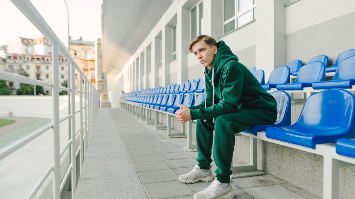Athletic young man in a green sports suit sits alone in the stands and looks away with a serious face. Young man resting sitting in the stands after training. Sports style.