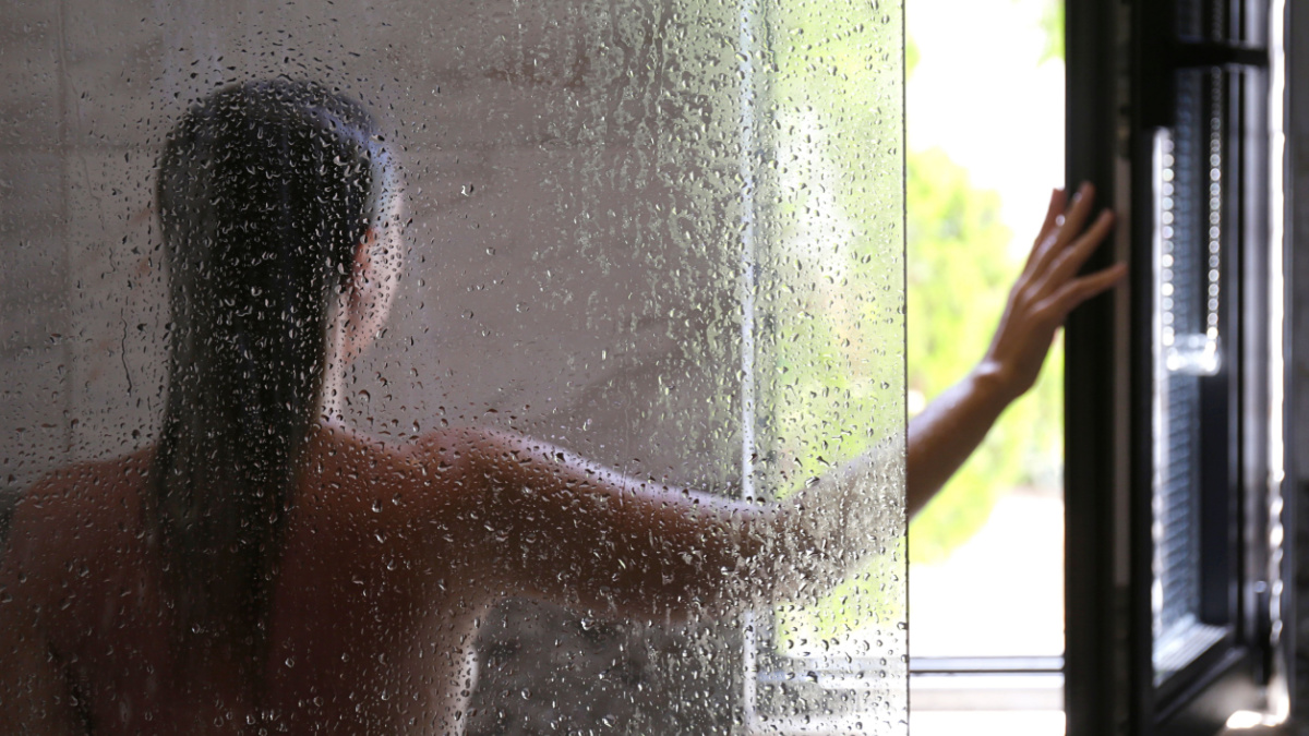 Back view of a naked woman in a shower cabin opening a window. Modern framless fixed glass panel, marble textured tile.