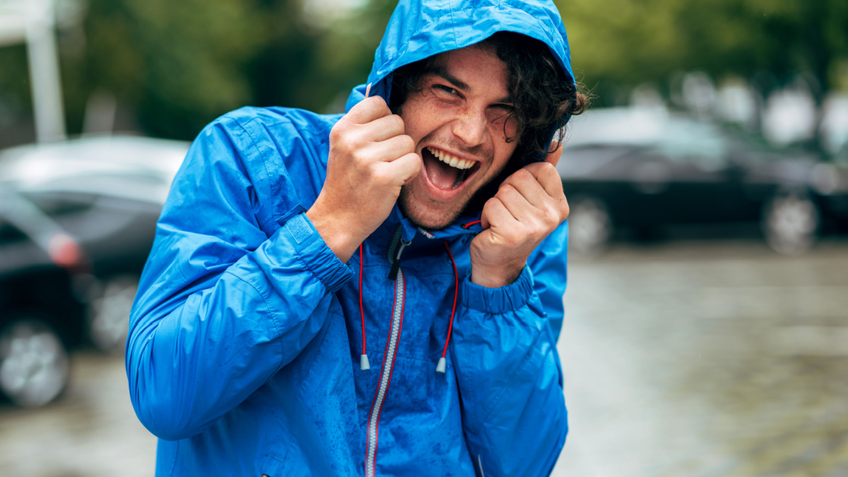 Happy man laughing and wearing blue raincoat during the rain outside. Handsome male in blue raincoat enjoying the rain in the city street. Male has joyful expression in rainy weather.