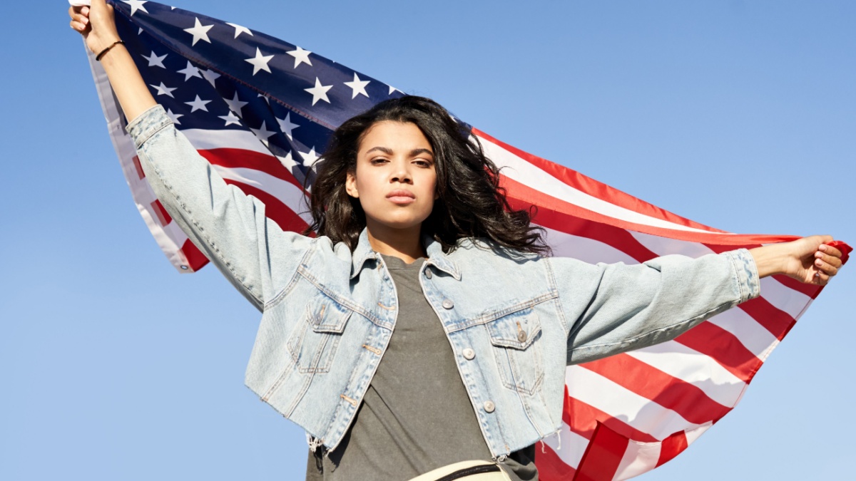 Proud serious young Afro American woman standing on street holding usa flag under blue sky looking at camera.