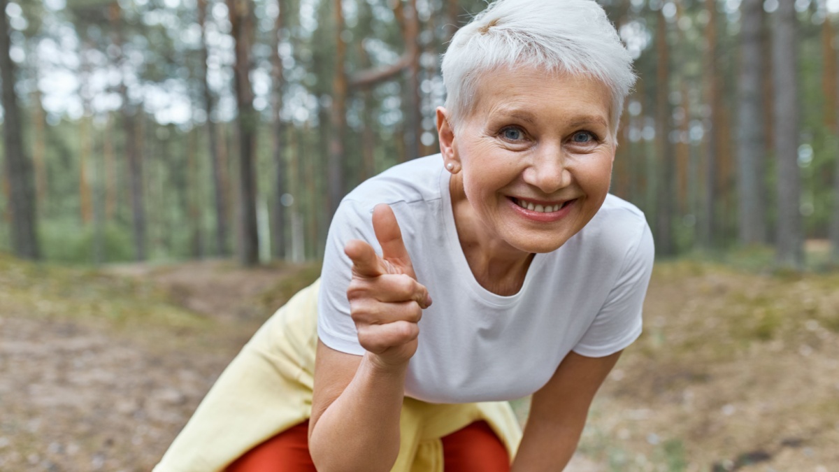 Outdoor view of energetic joyful female pensioner in sports clothes leaning forward, smiling and pointing fore finger at camera, inviting you to join her in morning run, having break during workout.