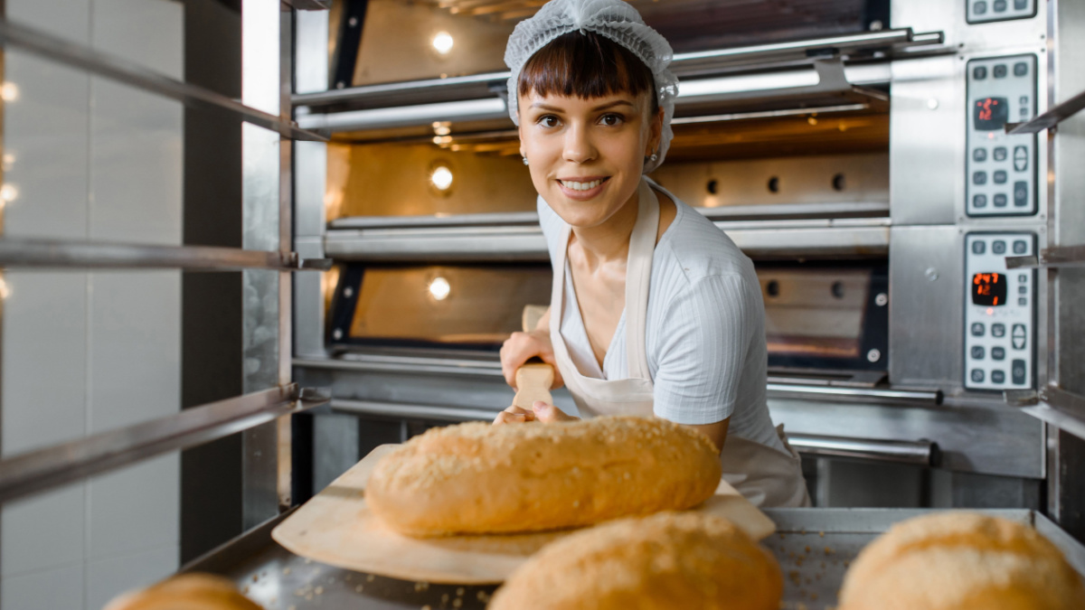 Close up of young happy smiling caucasian woman baker putting the fresh bread on the shelves/rack at baking manufacture factory. Tasty bread bakery