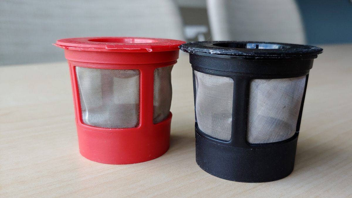 Reusable K cups for coffee machine. Filters are red and black with fine mesh. Individual coffee pods are sitting on a staff room table with chairs out of focus in the background.
