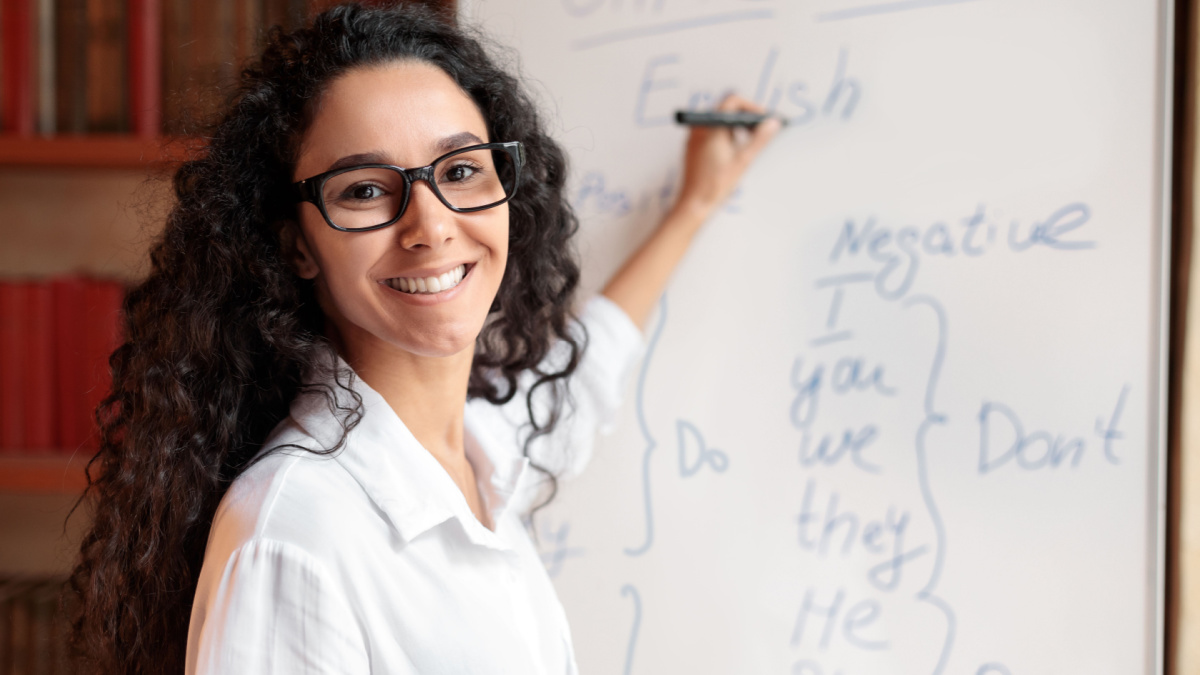 Education And Learning Concept. Portrait of smiling female teacher standing at whiteboard, explaining grammar rules to students. Excited woman in glasses looking at camera, writing on the board