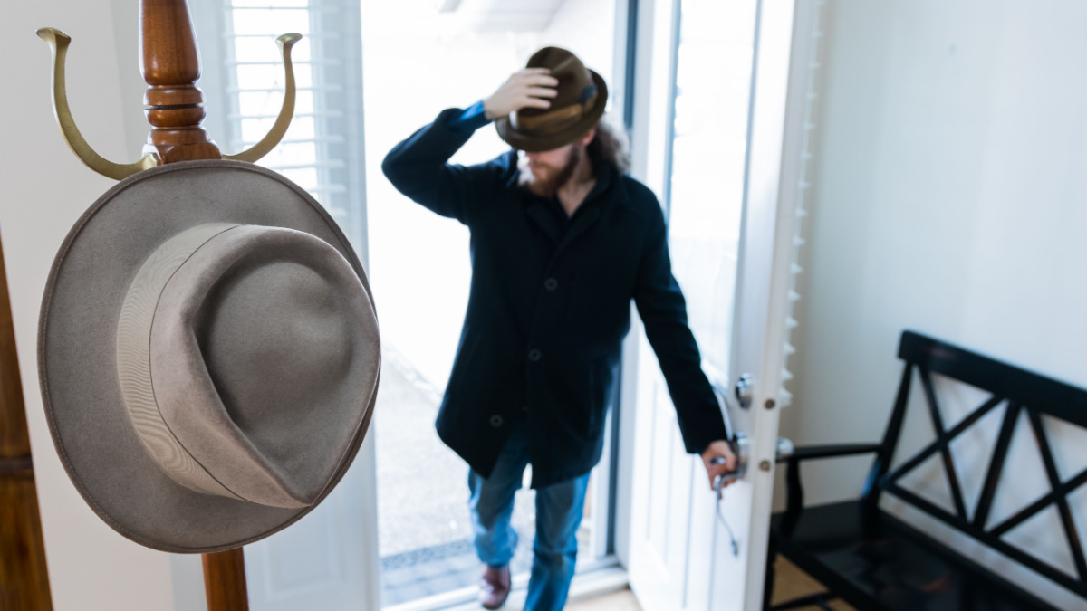 Coat rack at the front door with traditional hat. Man arriving home from work walking through front door wearing vintage fedora hat in background. Person in doorway to house entrance taking off hat