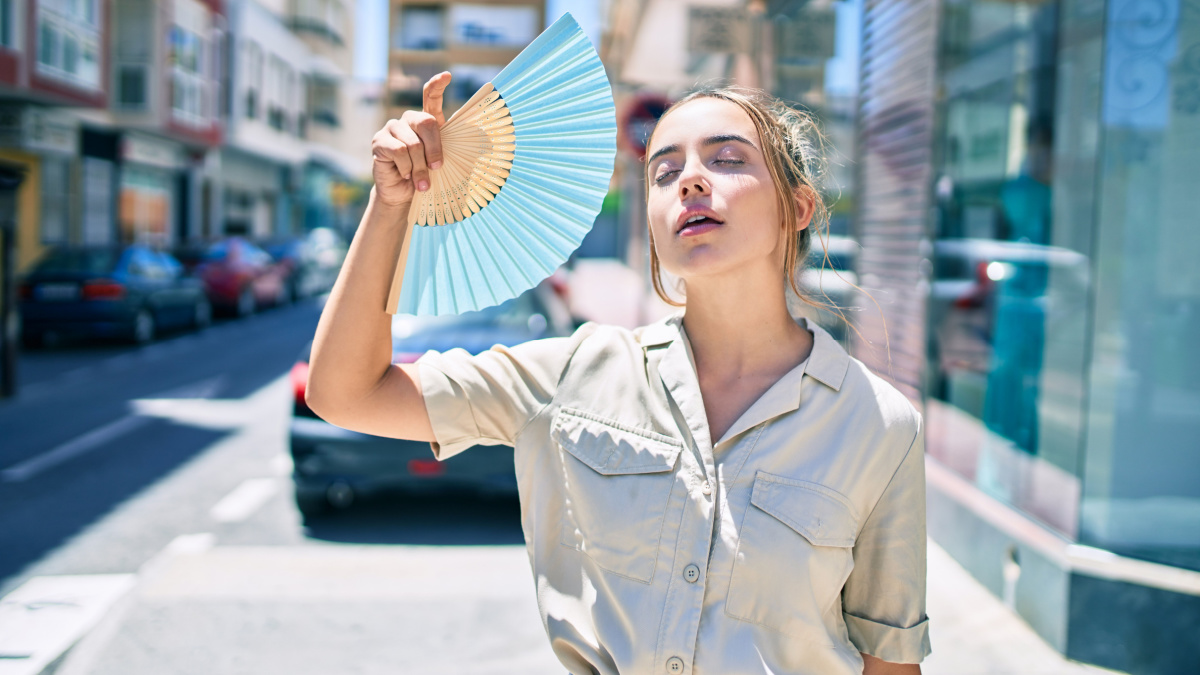 Young beautiful blonde caucasian woman smiling happy outdoors on a sunny day using handfan for hot weather