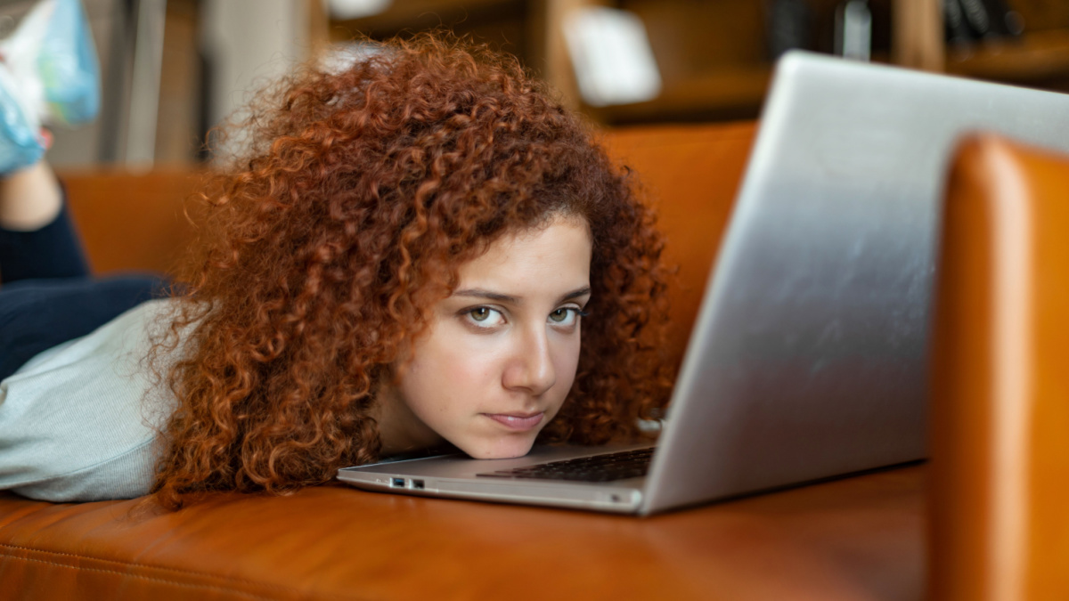 Sad bored young curly red-haired woman lying down on the couch in front of opened laptop and looks at the camera, feels melancholy and depression.