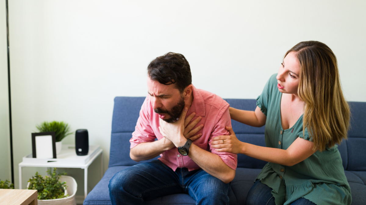 Adult latin man choking with a piece of food while sitting on the sofa with his young girlfriend at home