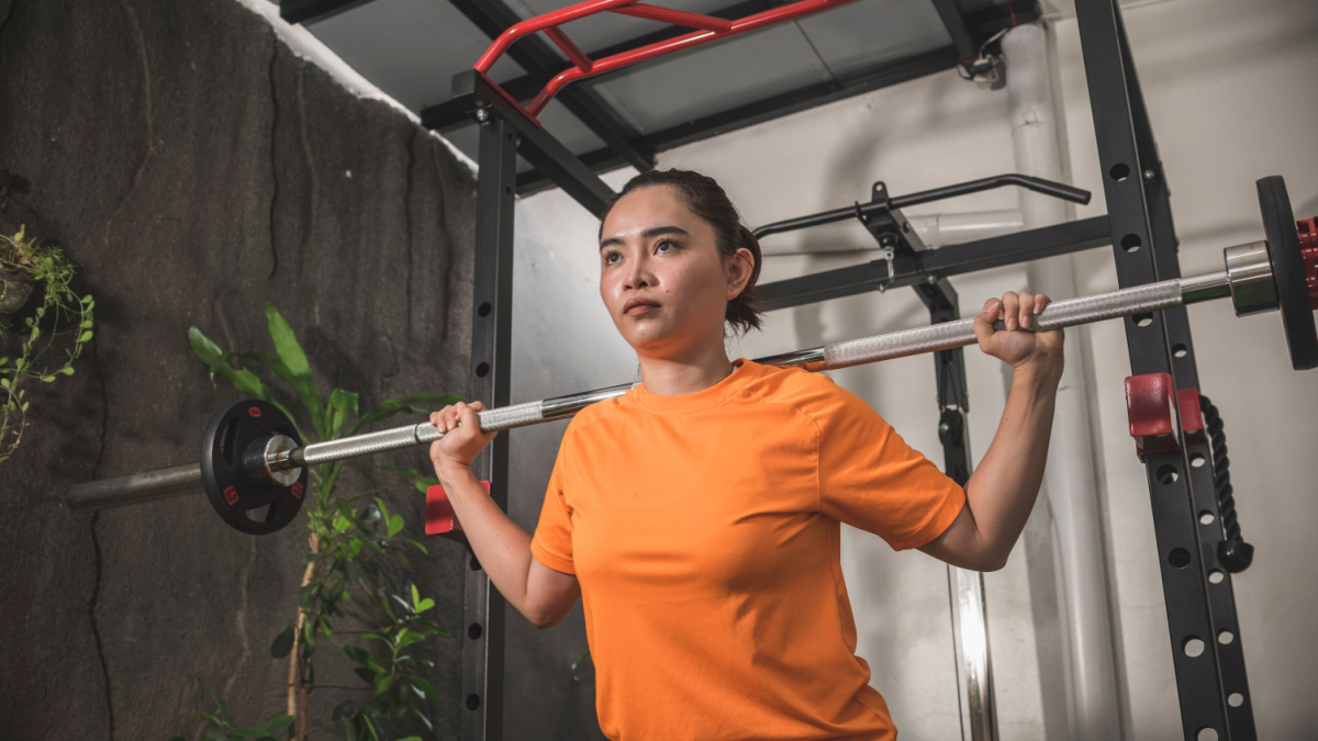 A determined millennial asian woman gets ready to do a set of barbell squats from a home workstation.