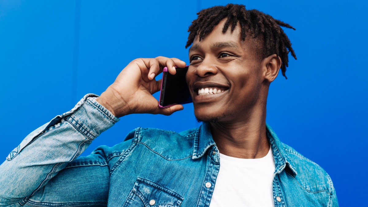 Happy curly african american man talking on cell phone, calling phone while walking outdoors against blue wall.