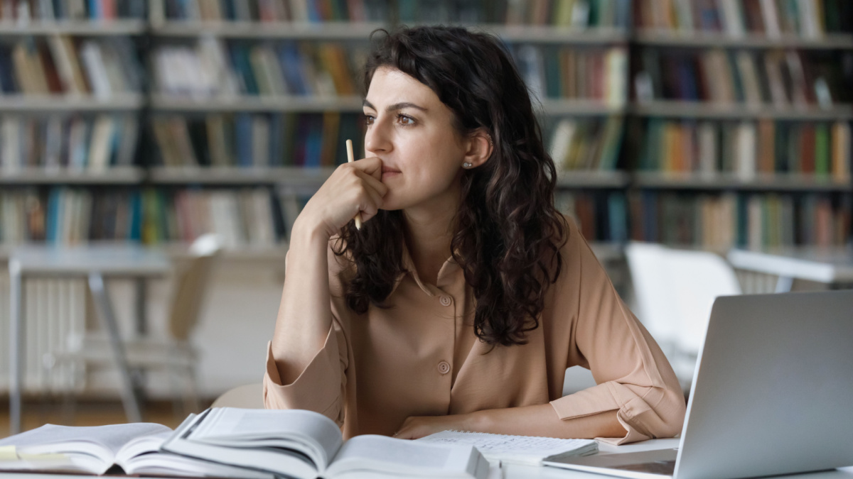 Young student girl sit at table with textbooks and laptop staring aside, studying alone in library, looks pensive and thoughtful search solution, prepare for exam, makes task feels confused or puzzled