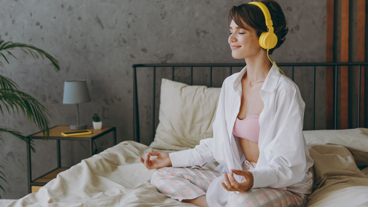 Young woman wear white shirt pajama headphones she lying in bed listen to music mantra meditating hold hands in yoga om gesture rest relax spend time in bedroom lounge home in own room hotel wake up