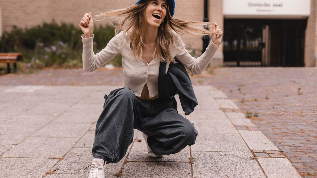 Full length radiant young caucasian girl kneels down against street. Blonde woman is wearing wide pants cargo, sweater top with buttons and blue panama hat, bag on shoulder. Girl look funky, fix hair.