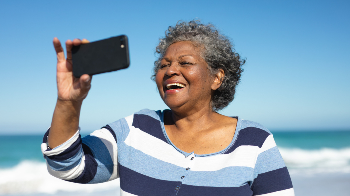 Front view close up of a senior African American woman standing on the beach with blue sky in the background, laughing and taking a selfie with a smartphone.