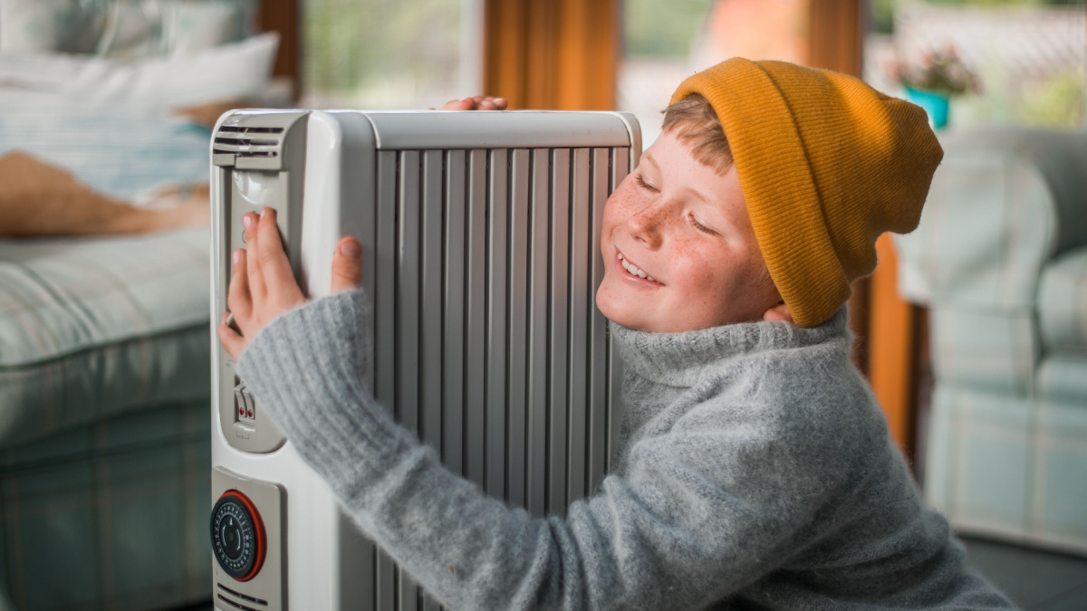 Funny child hugs the radiator. Kid in hat and warm clothes enjoying home heater. Heating of housing during the energy crisis in the winter cold season. Restrictions and savings of gas and electricity.