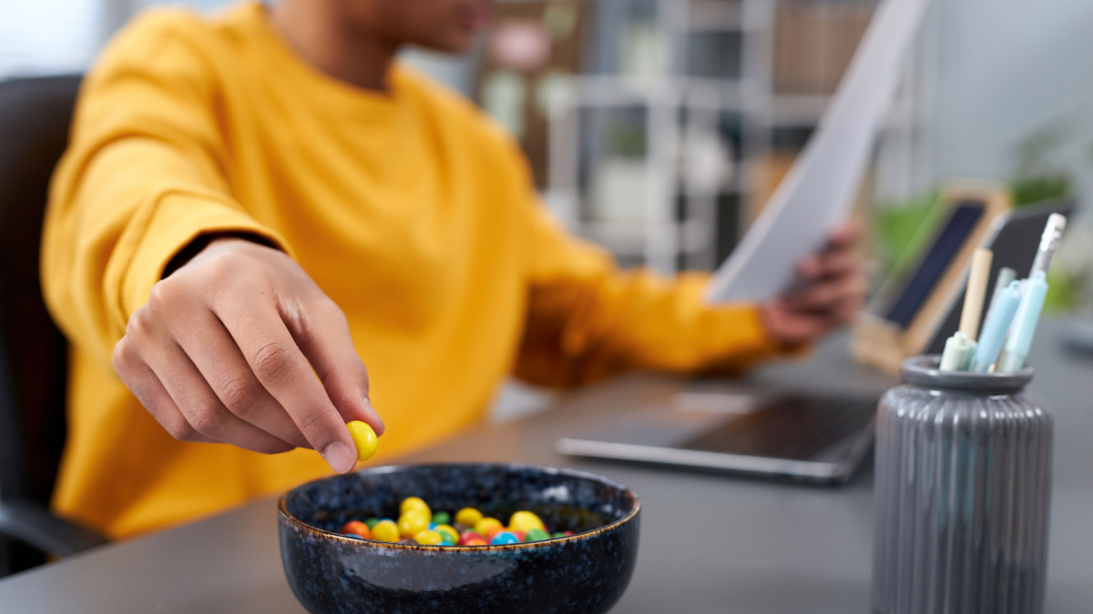 Close up of young man eating snacks while working or studying at home, focus on hand holding colorful candy, copy space