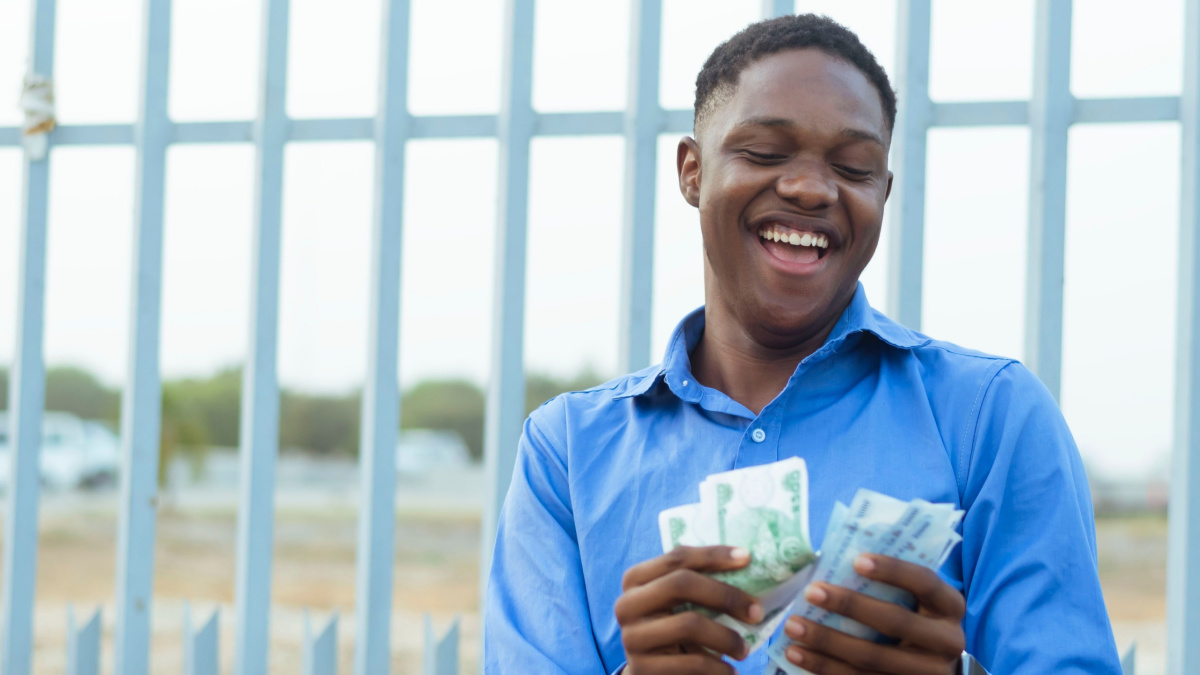 A cheerful young man in a blue shirt is happily counting a stack of cash bills in front of a tall metal fence, seemingly outdoors during the daytime.