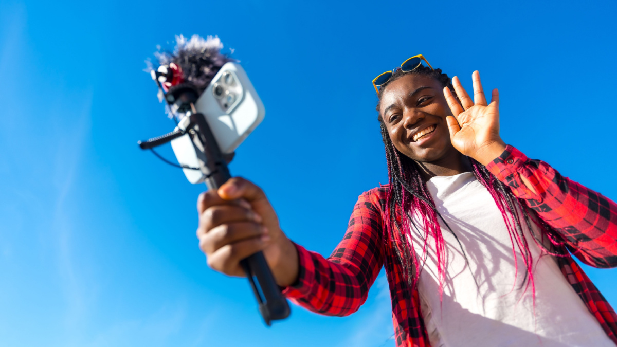 African woman waving while vlogging using mobile phone and microphone outdoors.