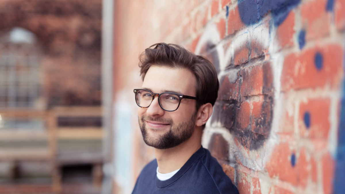 Portrait of Smiling Young Man with Facial Hair Wearing Eyeglasses and Leaning Against Brick Wall Painted with Graffiti.
