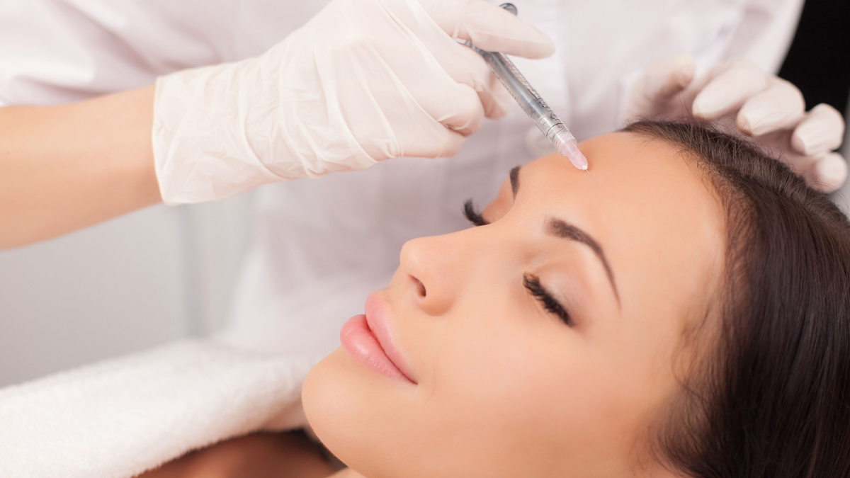 Close up of hands of expert beautician injecting botox in female forehead. The woman closed her eyes with joy. She is gently smiling