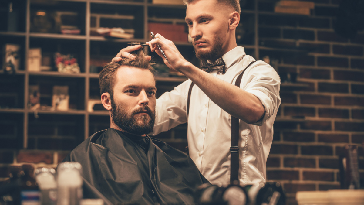 Making haircut look perfect. Young bearded man getting haircut by hairdresser while sitting in chair at barbershop