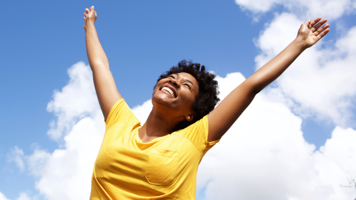 Portrait of cheerful young woman standing outside with her hands raised towards sky.