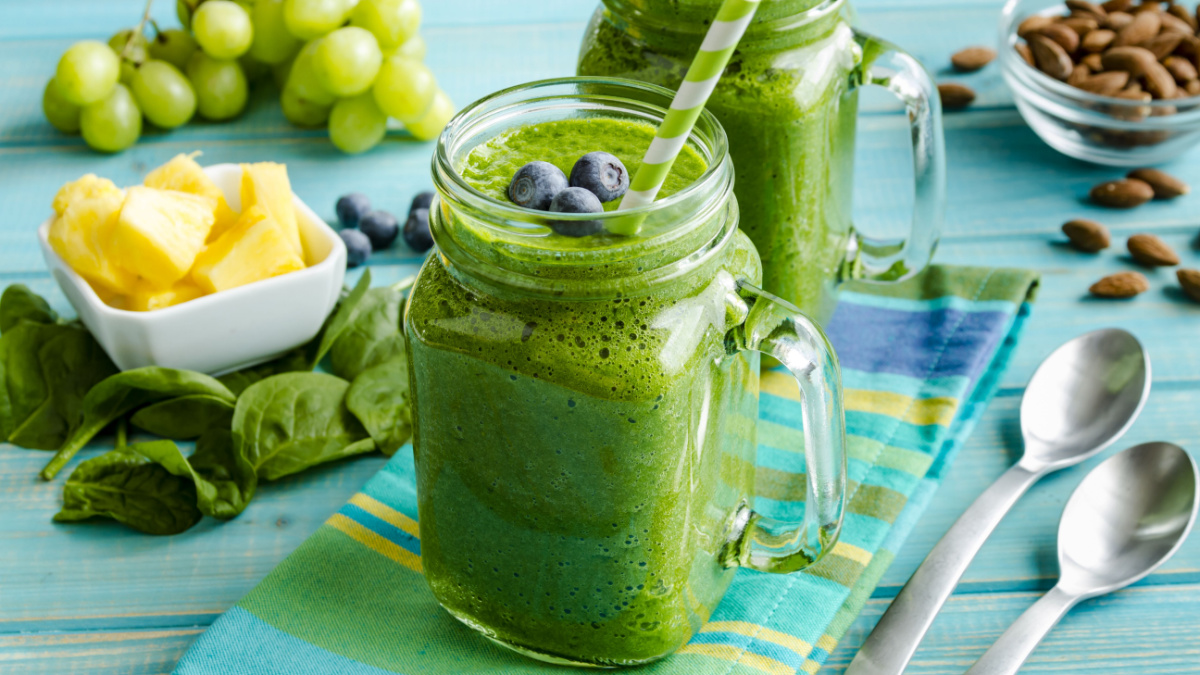 Mason jar mugs filled with green spinach and kale health smoothie with green swirled straw sitting with blue striped napkin and spoons