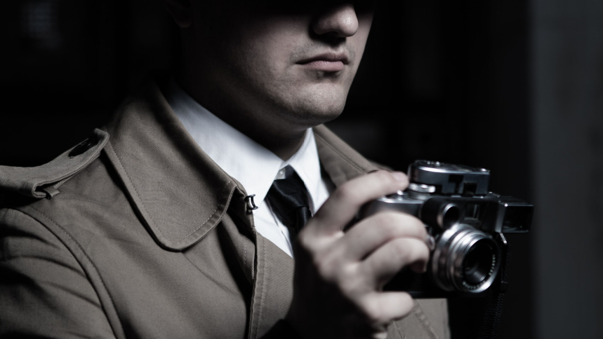 Young male spy agent wearing a hat, coat and a tie, holding rangefinder Leica film camera. Eyes hidden in the shadow.