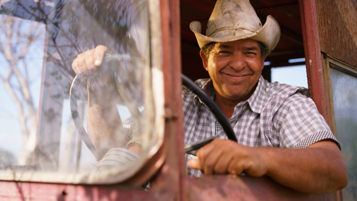 Farming and cultivations in Latin America. Portrait of middle aged hispanic farmer sitting proud in his tractor at sunset, holding the steering wheel. He looks at the camera and smiles happy.