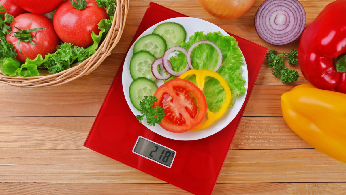 Plate with sliced fresh vegetables on digital kitchen scales over wooden background