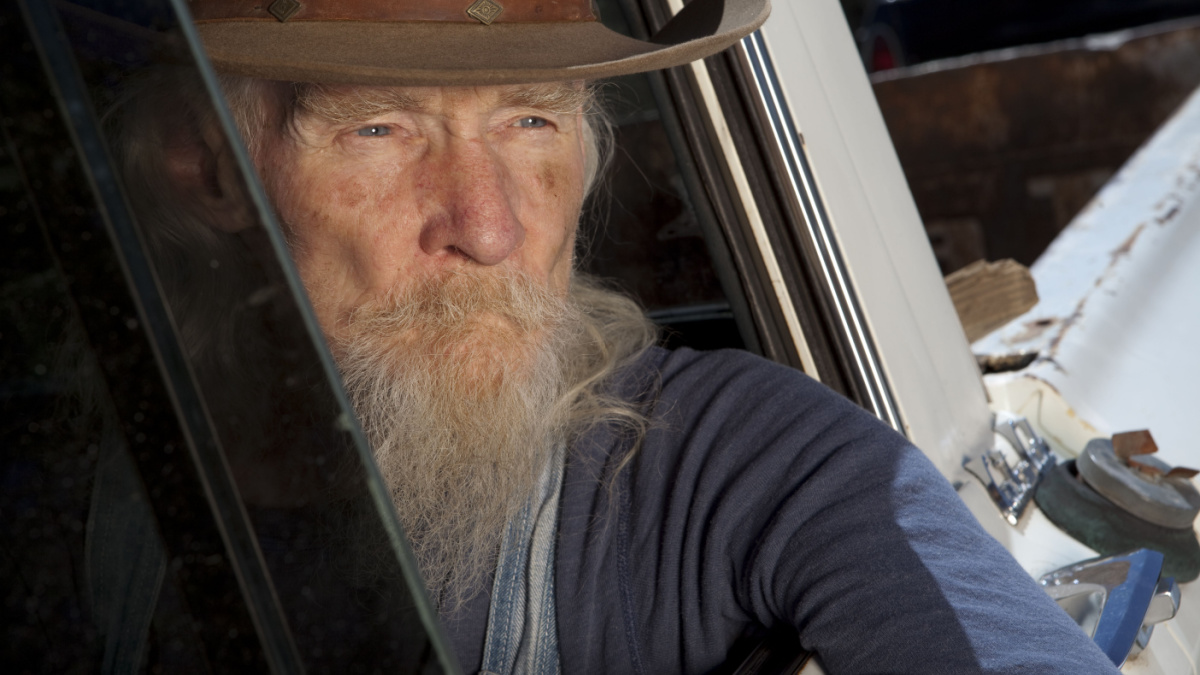 An elderly man with a white beard driving a pickup truck and staring out the window.