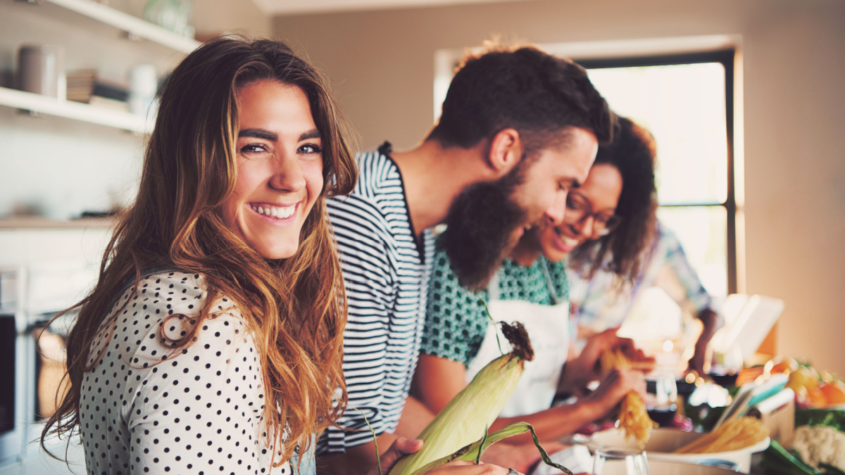 Laughing woman talking and preparing meals at table full of vegetables and pasta ready for cooking in kitchen.