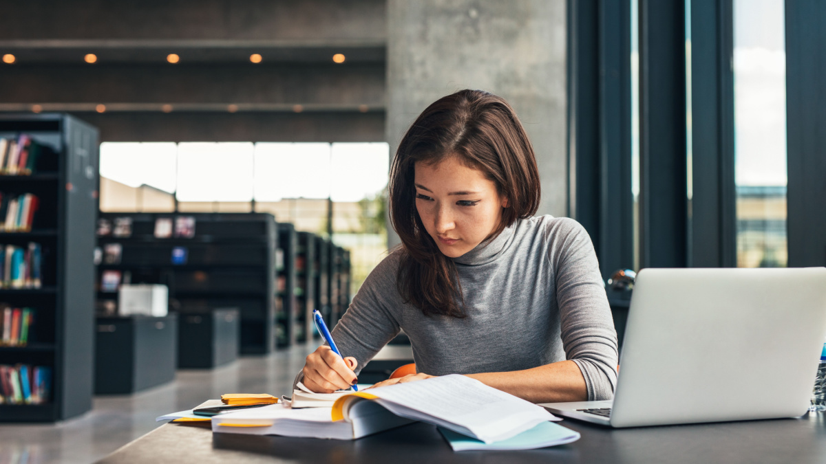 Female student taking notes from a book at library. Young asian woman sitting at table doing assignments in college library.