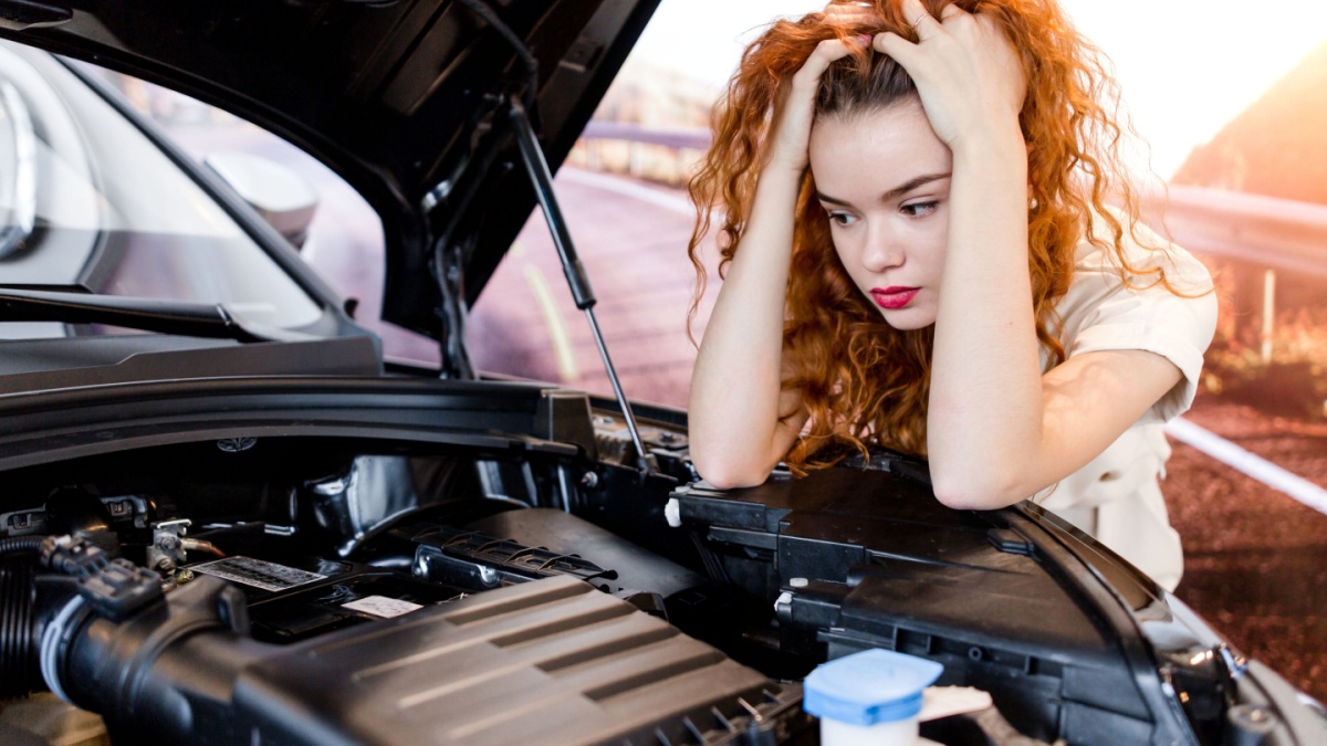 Redhead girl standing in front of broken car holding her head.